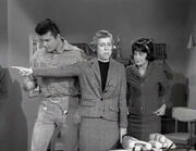 A black and white screenshot from the television series, The Beverly Hillbillies shows Max Baer Jr. as Jethro, Nancy Kulp as Jane Hathaway and Sharon Tate as Janet Trego, a secretary. Tate is wearing a business suit and a dark wig and is watching Miss Hathaway
