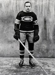 Full profile of a balding ice hockey player in full uniform leaning forward on his stick with a serious look on his face.