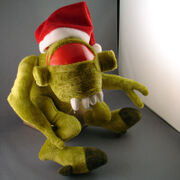 A green plush toy of a Vortigaunt sits against a gray wall. The toy is wearing a Santa Claus hat, leaving only the dominant red eye visible.