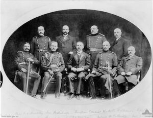 Formal group portrait of nine men, four sitting at the front and five standing behind. Three are wearing suits; the others are wearing formal double breasted military uniforms with sashes.